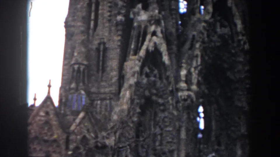 1959 Barcelona 8mm frame of Sagrada Familia towers and ornate facade elements, with pale daylight sky visible between vertical forms.