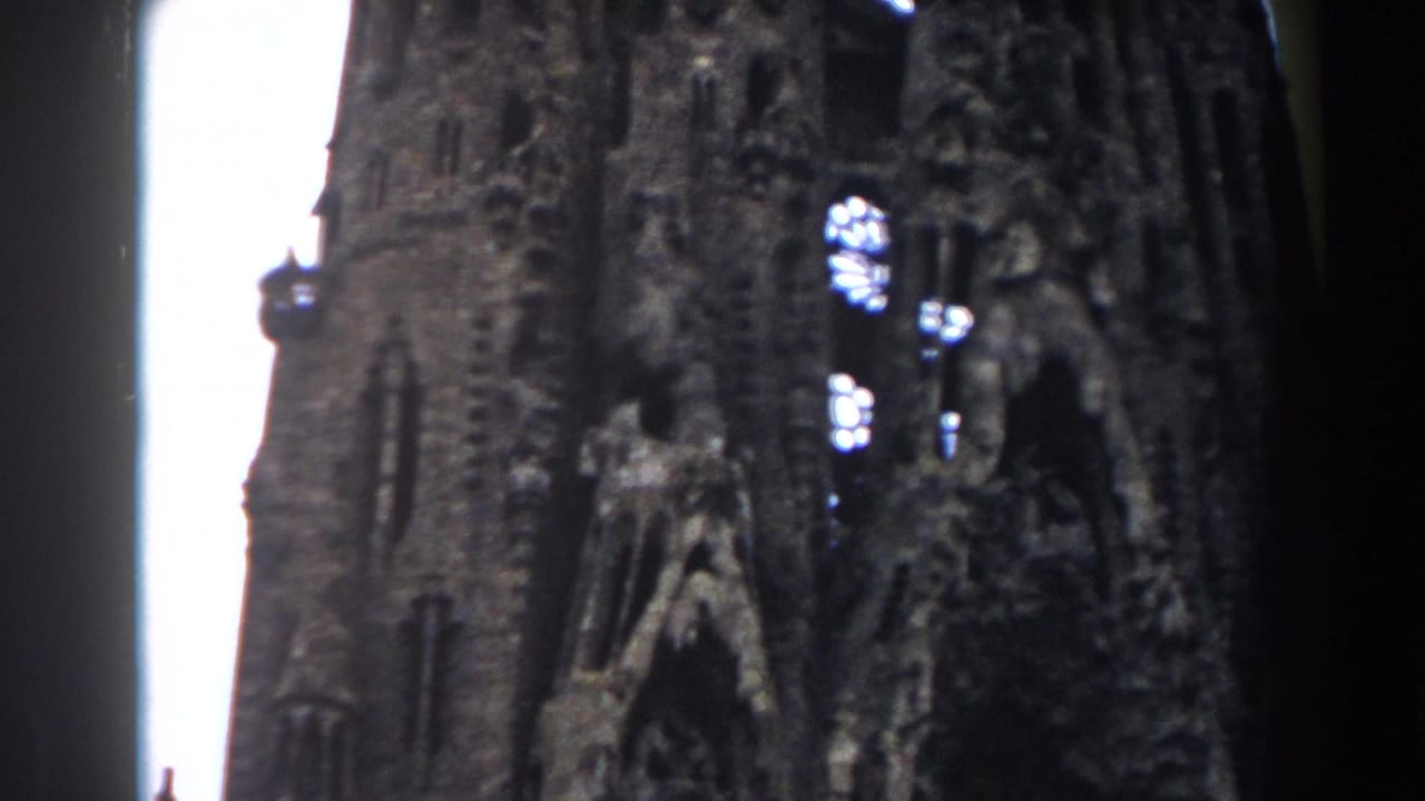 1959 Barcelona home-movie frame looking up at Sagrada Familia spires with carved stonework and bright sky behind them.