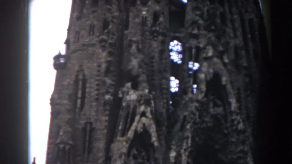 1959 Barcelona home-movie frame looking up at Sagrada Familia spires with carved stonework and bright sky behind them.