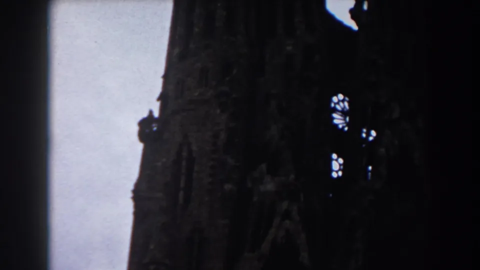 1959 Barcelona frame showing a close upward view of Sagrada Familia stonework, bright window openings, and pale sky.