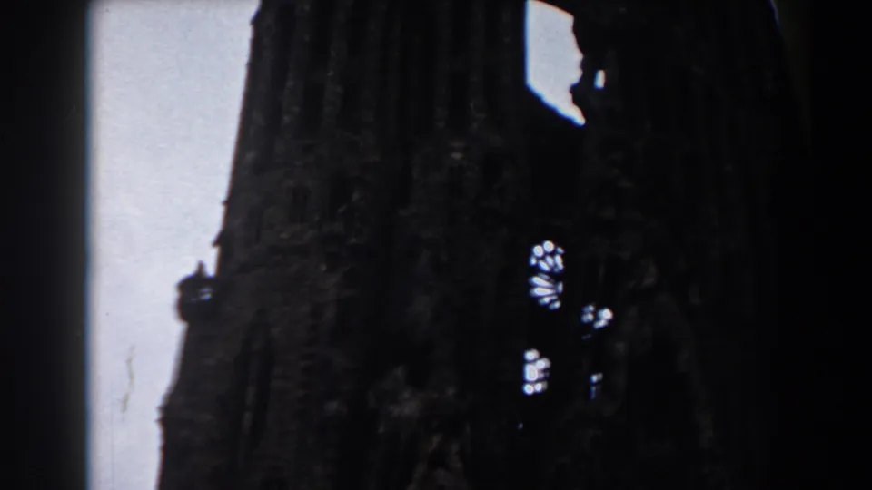 1959 Barcelona home-movie frame of a shadowed Sagrada Familia spire, small bright window patterns, and pale sky along the left edge.
