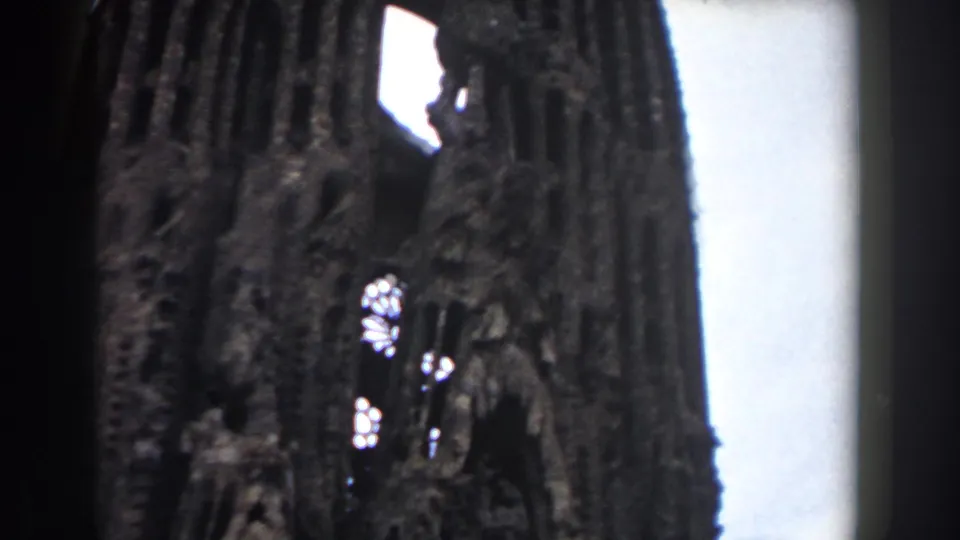 1959 Barcelona frame looking upward at a tall Sagrada Familia tower, with dark carved stone, bright openings, and pale sky to the right.