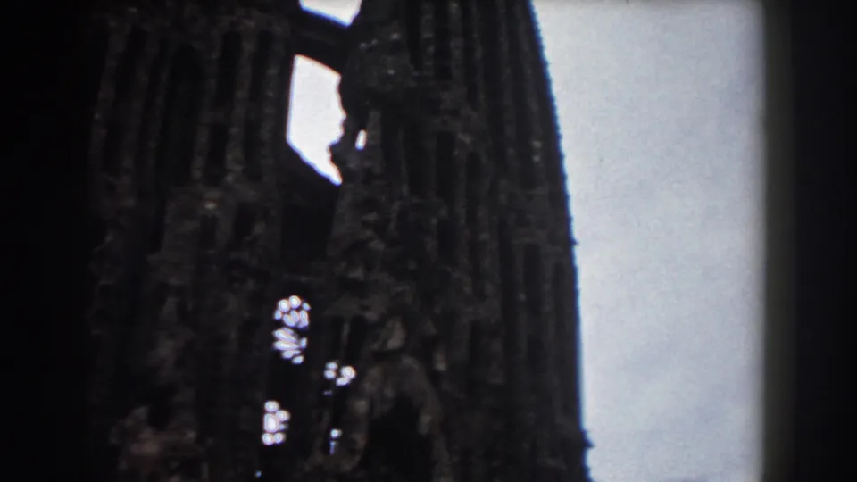 1959 Barcelona 8mm frame showing dark Gothic church or cathedral towers with damaged openings and pale sky behind them.