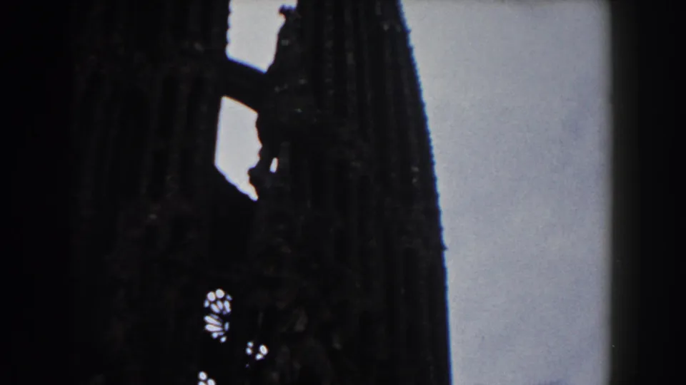 Shadowed 8mm view of a Barcelona cathedral or church facade, with tall stone towers and small bright openings visible through the structure.