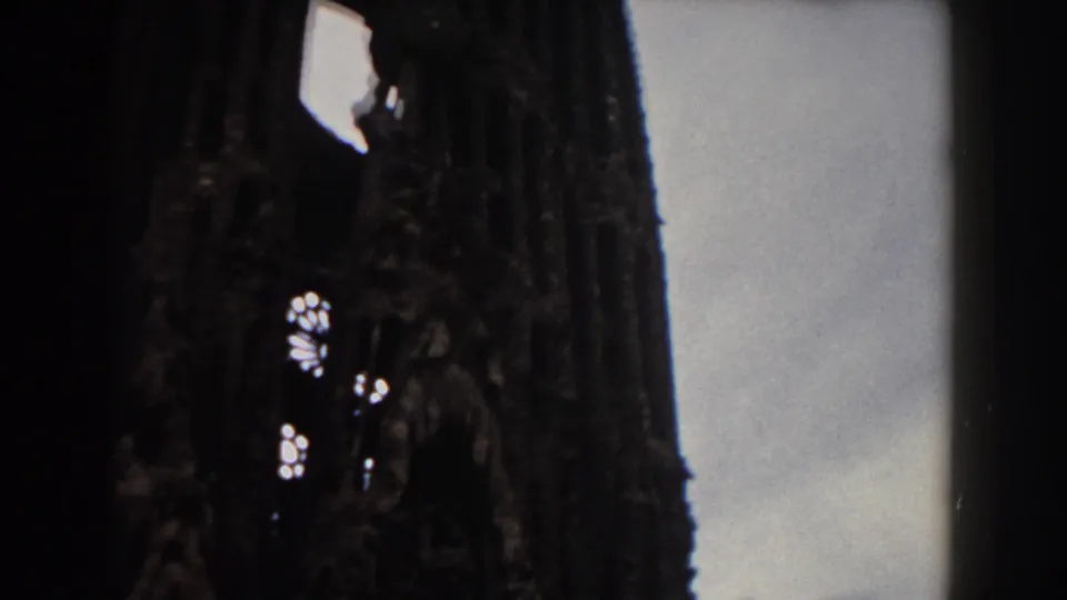 Mid-century 8mm frame of Sagrada Familia's dark facade and spires from the square in Barcelona.