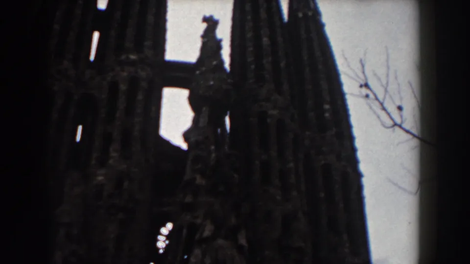1959 Barcelona 8mm frame showing Sagrada Familia spires, pale sky, and a bare tree branch at the edge.