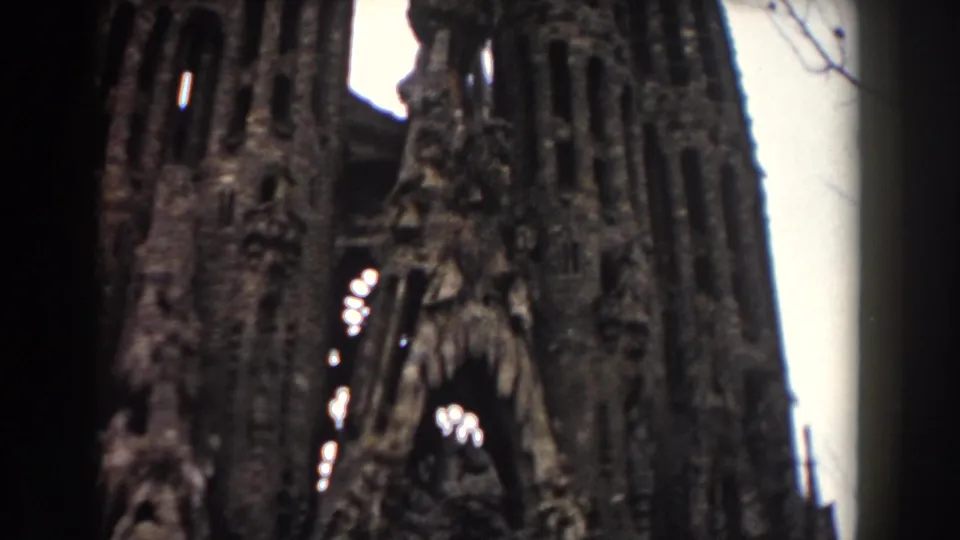 1959 Barcelona home-movie frame with the camera aimed up at an ornate stone facade, arched recesses, and a dark vertical shadow.