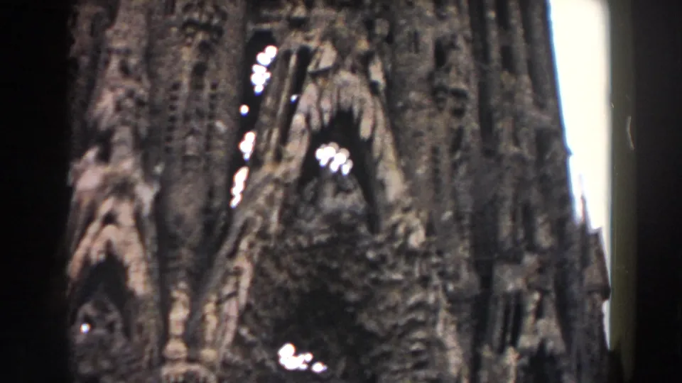 1959 Barcelona frame of a monumental stone facade with an arched opening, clustered carvings, and handheld 8mm softness.