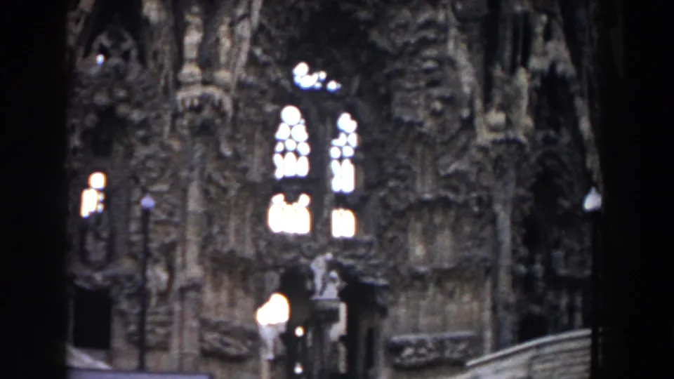 1959 Barcelona 8mm frame showing part of the Sagrada Familia facade, bright arched windows, lamps, steps, and a small visitor figure.