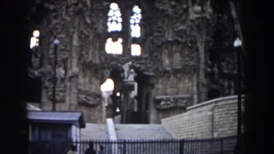 1959 Barcelona view of the Sagrada Familia exterior with stained-glass-like window openings, railings, steps, lamps, and a visitor at the entrance.