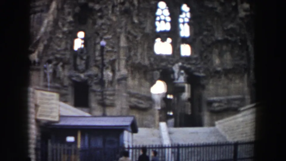 1959 Barcelona home-movie frame showing the Sagrada Familia lower facade, a small kiosk, iron fence, steps, and visitors partly visible below.