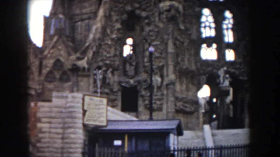 1959 Barcelona frame of the Sagrada Familia exterior with a kiosk, posted signs, iron fence, lamps, steps, and a visitor beside the entrance.