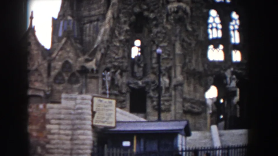 1959 Barcelona home-movie frame showing an ornate Gothic church facade with arched windows, carved stone figures, a small sign, and a lamppost.