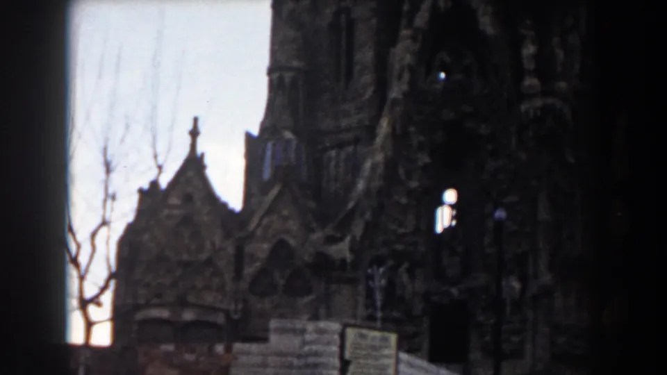 1959 Barcelona frame with a church gable, partial tower, arched details, foreground sign and fence, brickwork, and a bare tree at left.