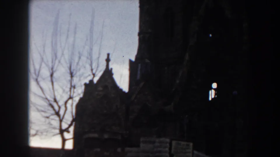 Final 1959 Barcelona frame showing the church facade in darker exposure, with a gable silhouette, bare branches, foreground sign, and arched light openings.