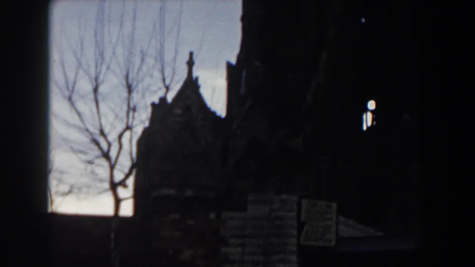 1959 Barcelona home-movie frame showing a shadowed Gothic exterior and leafless branches against a fading sky.