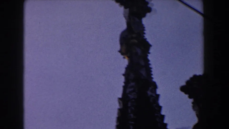 1959 Barcelona home-movie frame focusing on a single dark Gothic spire beneath a pale blue sky.