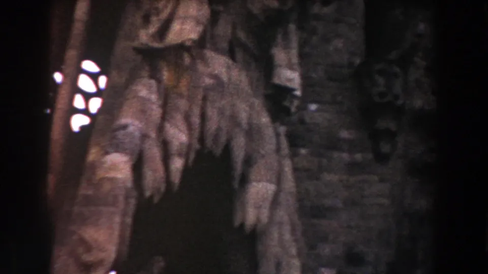 Dim 8mm view of rough stone carvings and a narrow stained-glass-like opening inside a Barcelona historic interior.