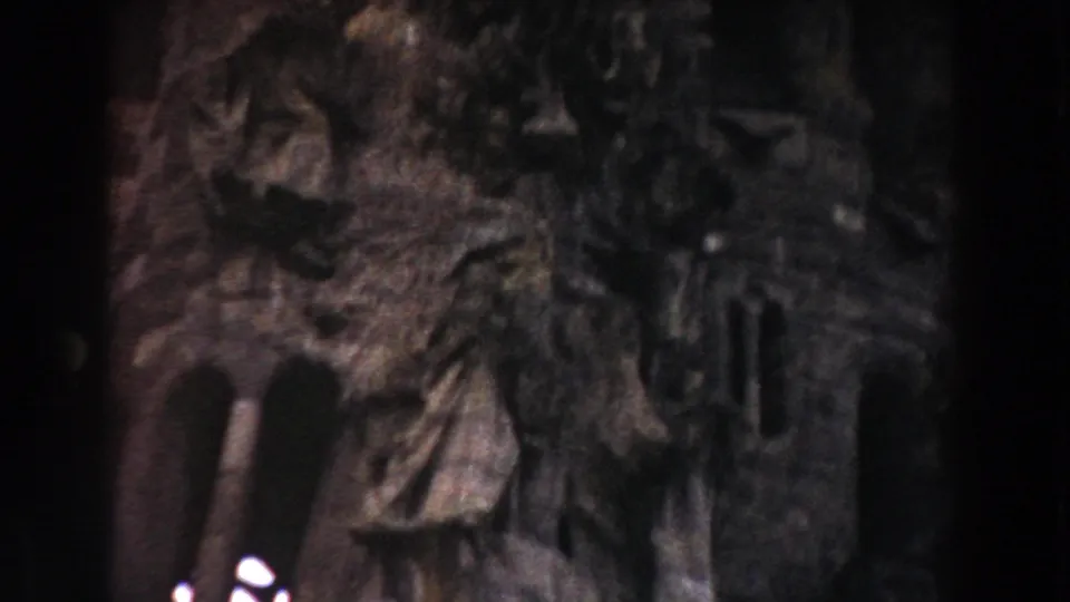 1959 Barcelona home-movie frame with clustered stone carvings, masonry texture, and a small sculpted figure in shadow.