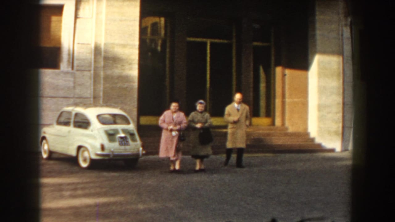 In 1959 Barcelona, three adults in period coats are seen near a parked white compact car outside a broad building facade.