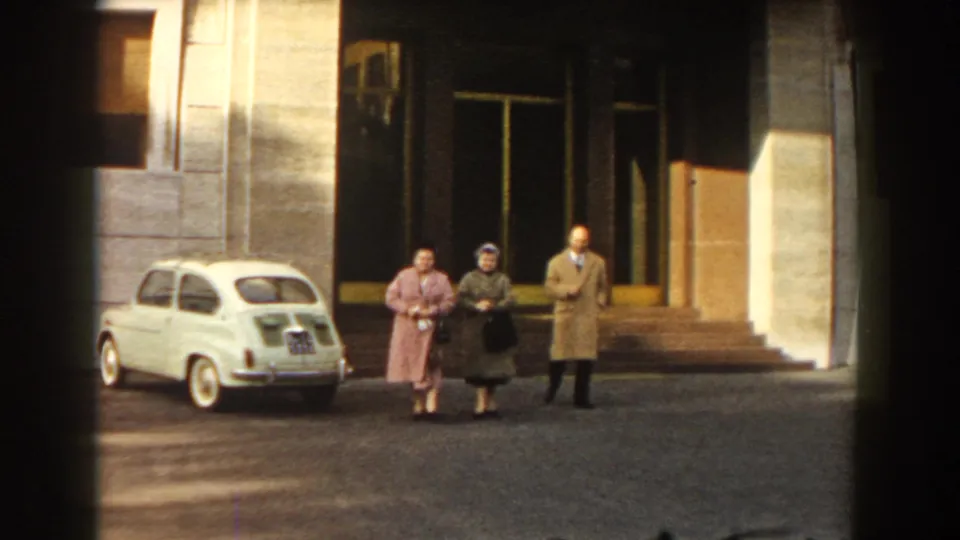 In 1959 Barcelona, three adults in period coats are seen near a parked white compact car outside a broad building facade.