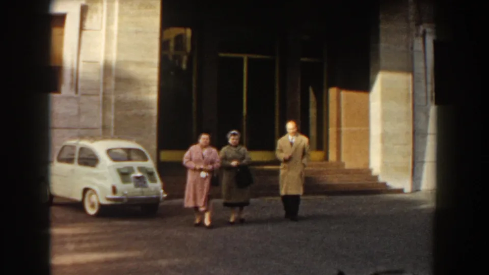 Three adults in 1950s dress walk together in Barcelona beside a parked white Fiat 500 and a large building entrance.