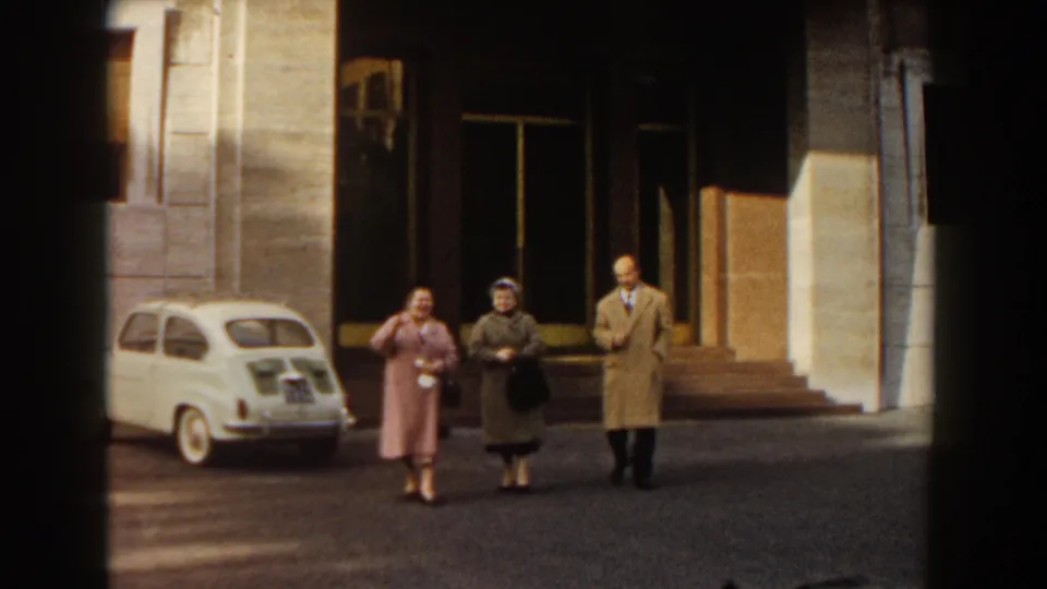 Two women and a man walk toward the camera in a 1959 Barcelona street scene, with a white compact car behind them.