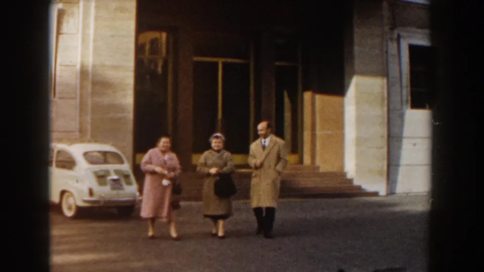The group pauses near a building entrance in Barcelona, with the white compact car partly visible at the left edge.
