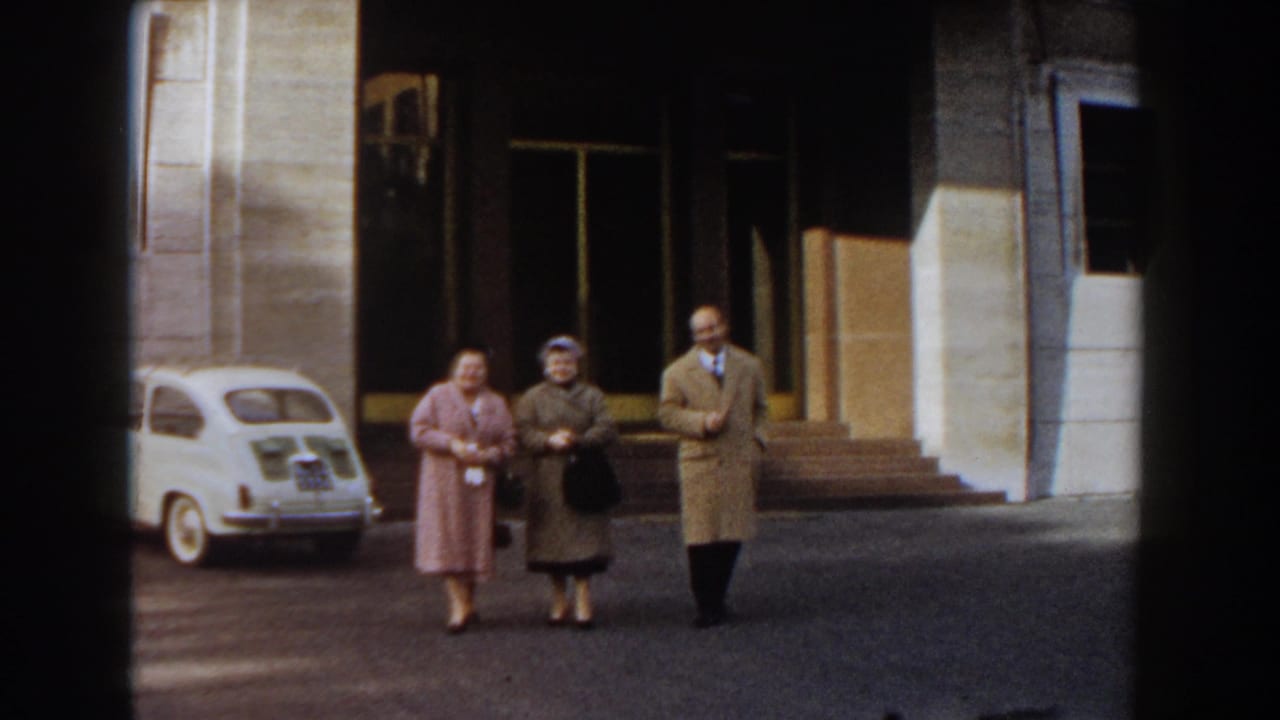 Three adults in coats walk near a white compact car outside a stone building entrance.