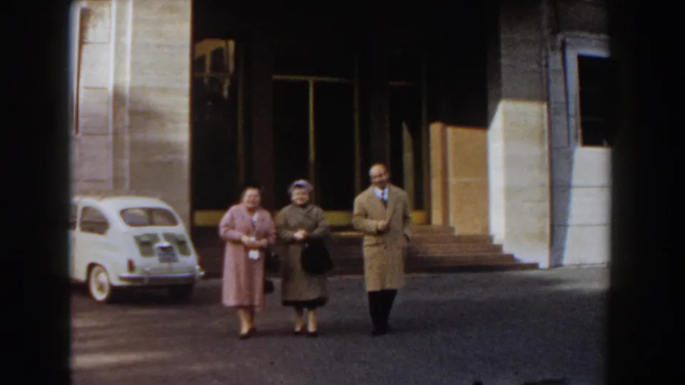 In 1959 Barcelona, two mature women and a man in coats walk toward the camera beside a white compact car and tall building doors.