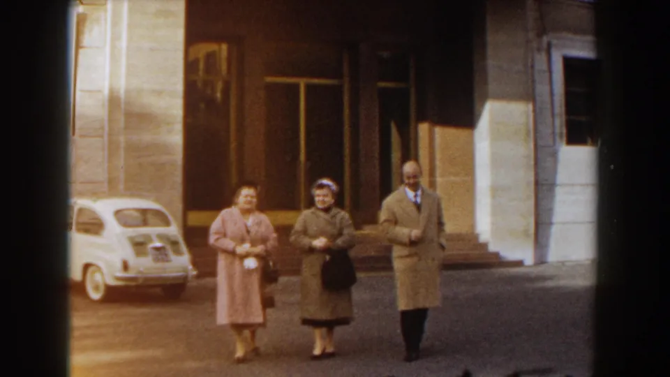 The adults approach the camera on a paved Barcelona street, with the white compact car and large building entrance remaining visible behind them.