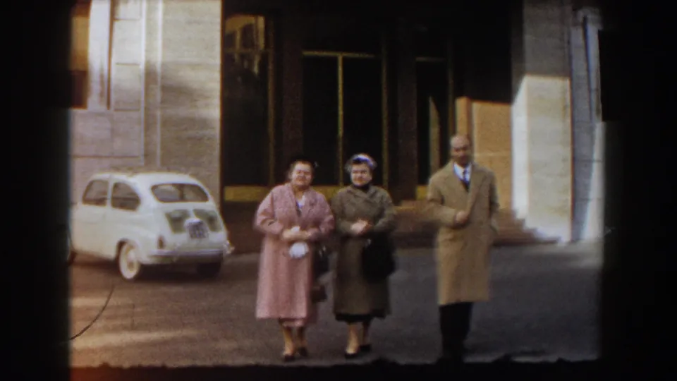 Near the end of the 1959 Barcelona clip, the two women and man pass close to the camera with the white car and building entrance behind them.