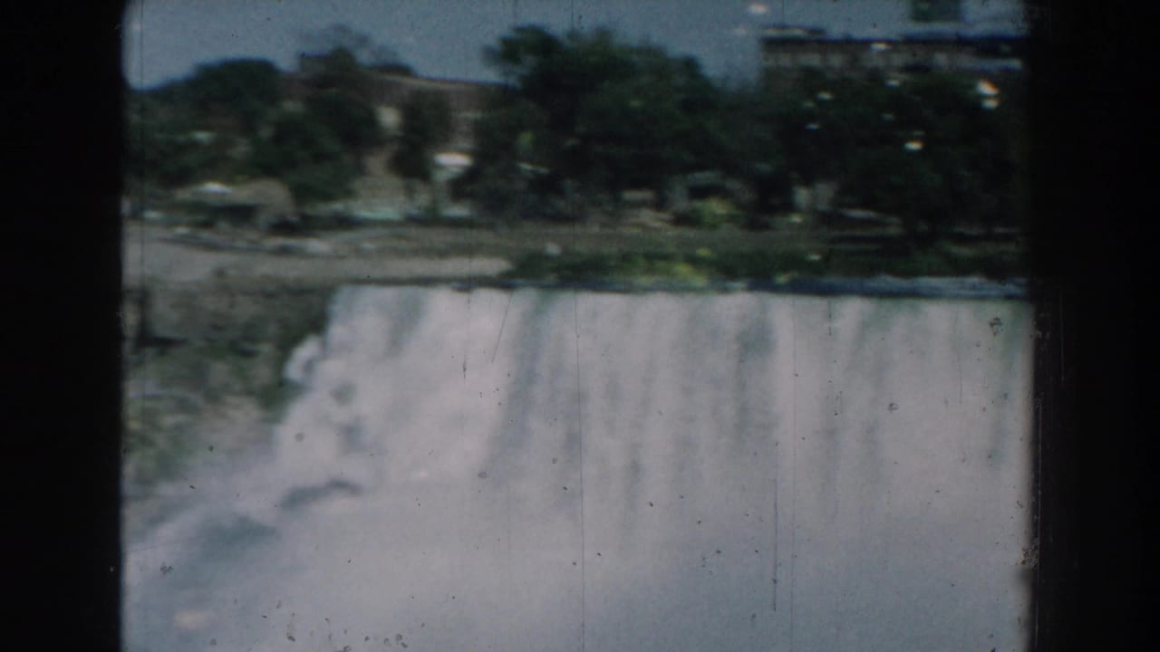Water pours over a wide dam with trees and buildings in the background.