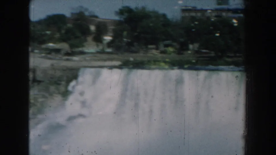 1959 Michigan 8mm frame showing a broad dam spillway, misting water, tree-lined bank, and distant urban buildings.
