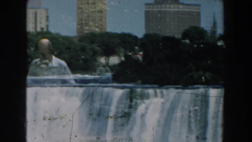 1959 Michigan home-movie frame with a light-shirted man near a dam overlook, rushing water, trees, and city buildings behind him.