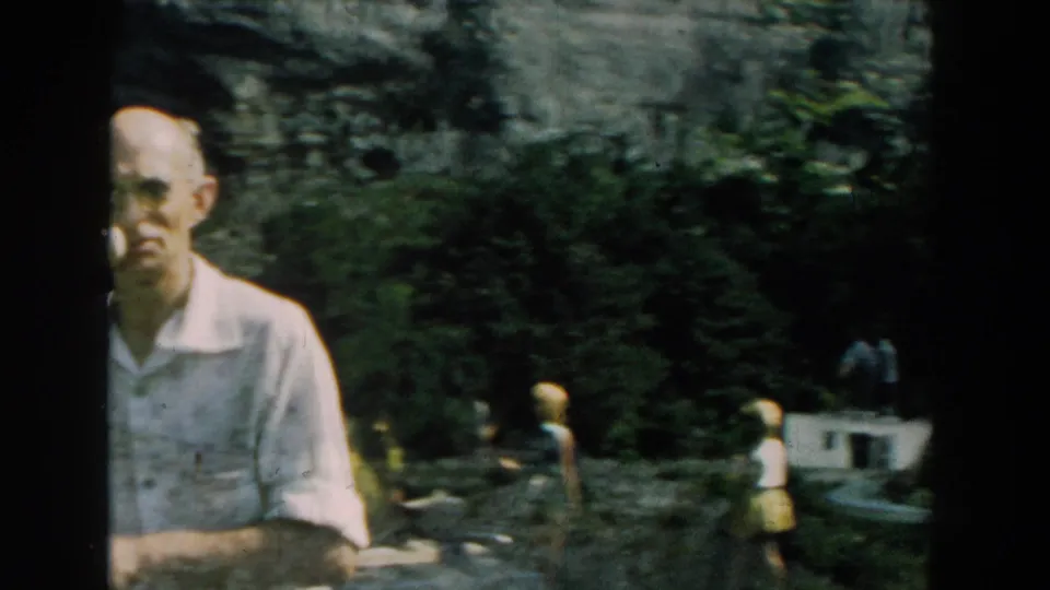 A 1959 Michigan 8mm frame shows an adult man near the camera, with shrubs, rockwork, visitors, and the dam area behind him.