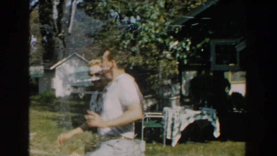 Adult man in a light sleeveless shirt moves across a Michigan backyard with a small house, trees, and patio items behind him.