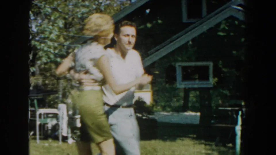 Adult couple dances on a sunlit Michigan lawn, with the woman in a light top and skirt and the man in a sleeveless shirt and trousers.