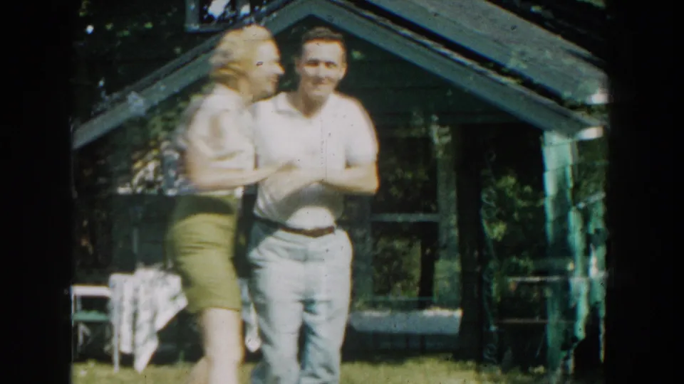 The couple continues dancing in the backyard, with the man facing the camera and the woman beside him near chairs, trees, and a small house.