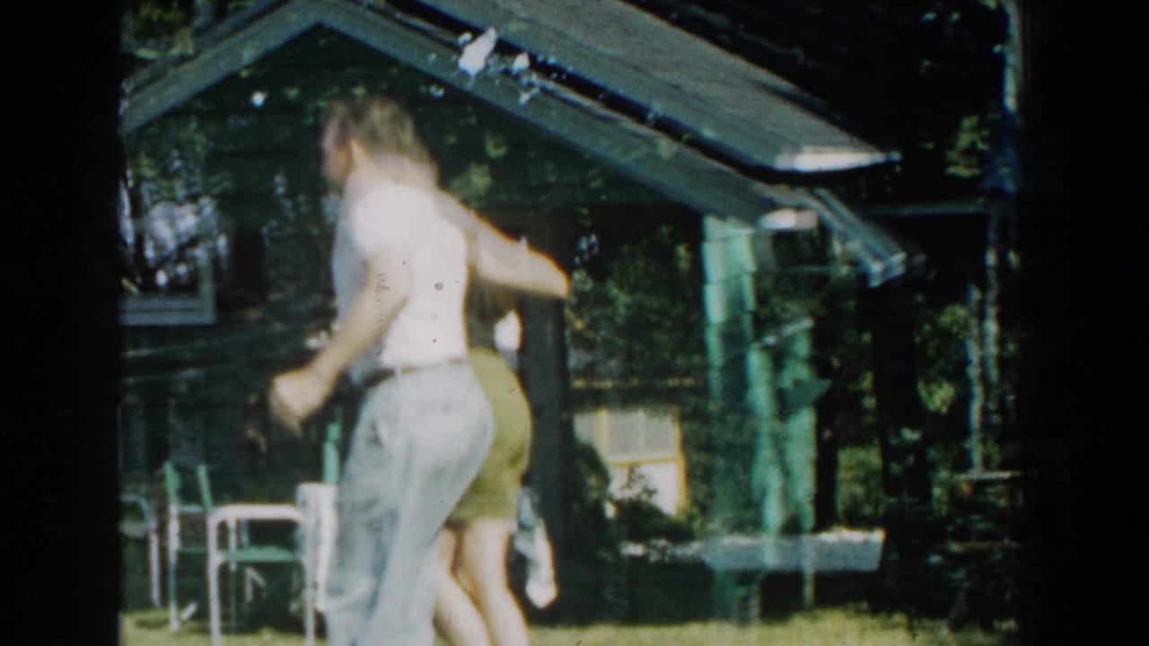 Man and woman dancing on grass beside a small cabin in a wooded yard.
