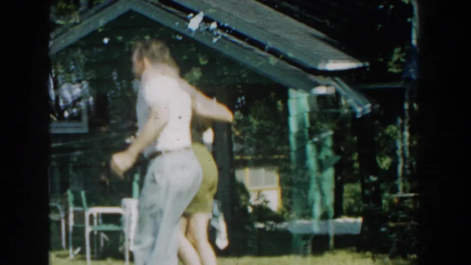 In Michigan 1959 home-movie footage, a man and woman move together on the lawn beside a green cabin with trees and yard furniture nearby.