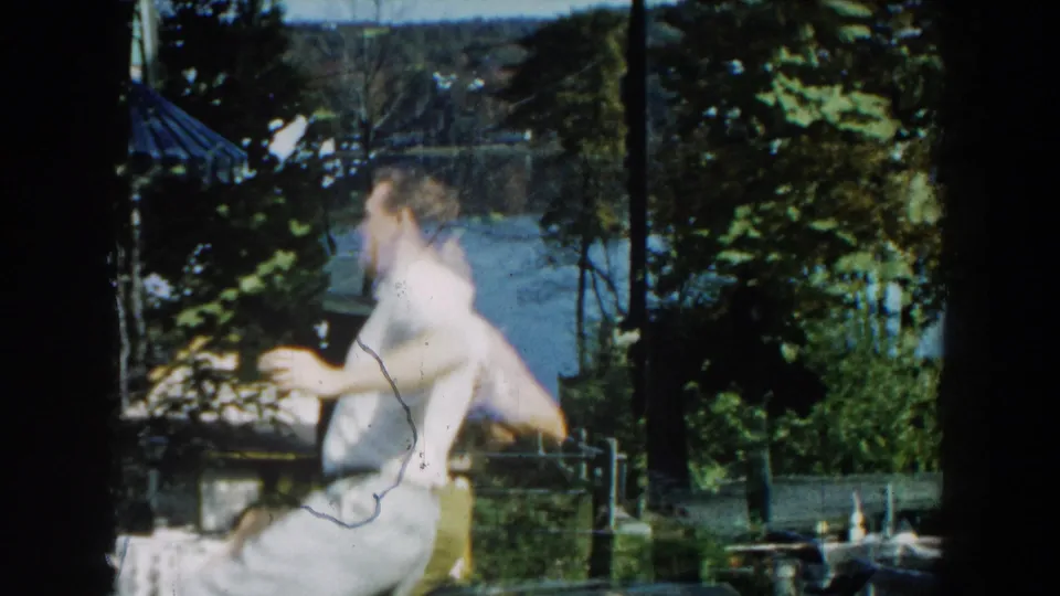 A man moves across the lawn during the backyard dance, partially blurred by motion, with trees, lake water, and yard furniture behind him.