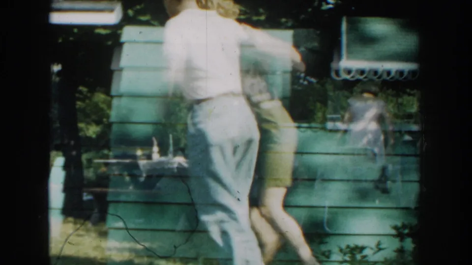 The couple passes near the green cabin wall during the dance, with a table, chairs, and shaded yard details visible around them.