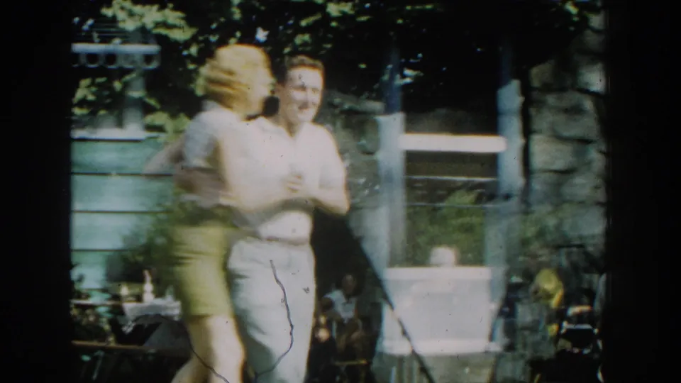 The woman turns quickly while the man faces her near the green-sided house, with trees and a window behind them.