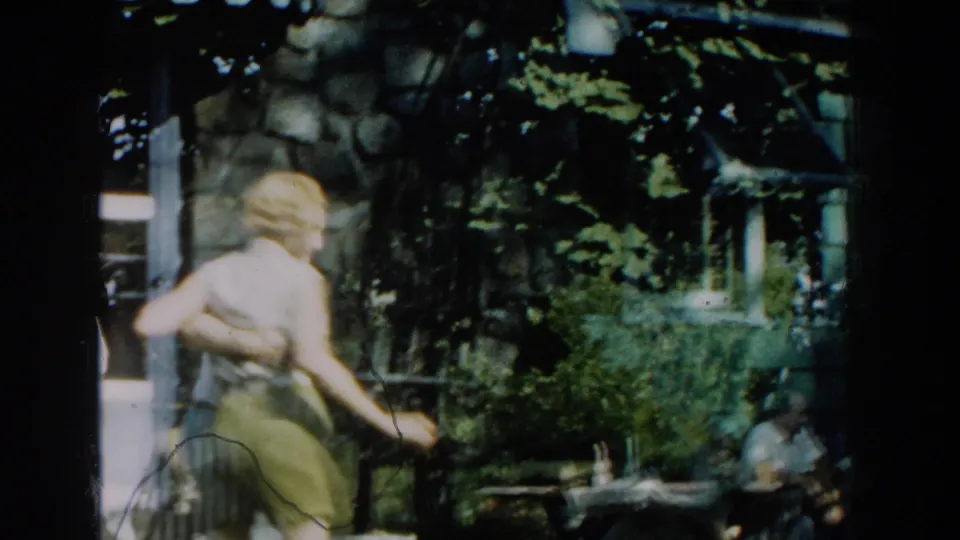 The woman turns away from the camera during the dance, with a stone wall, leafy trees, and shaded outdoor furniture visible nearby.