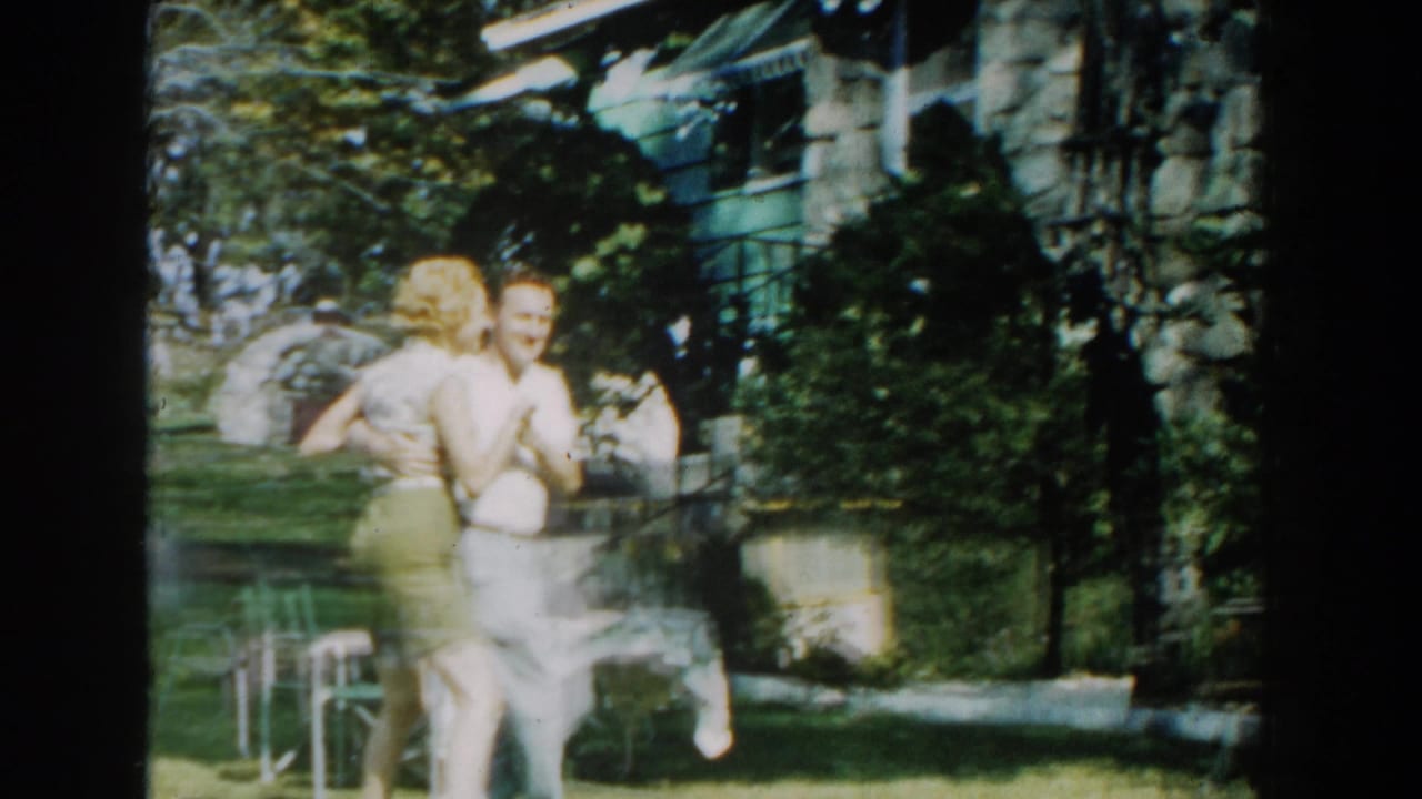 In 1959 Michigan home-movie footage, a couple dances beside a green-sided house as a nearby onlooker watches from the yard.