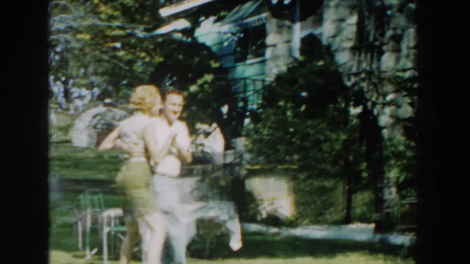 In 1959 Michigan home-movie footage, a couple dances beside a green-sided house as a nearby onlooker watches from the yard.
