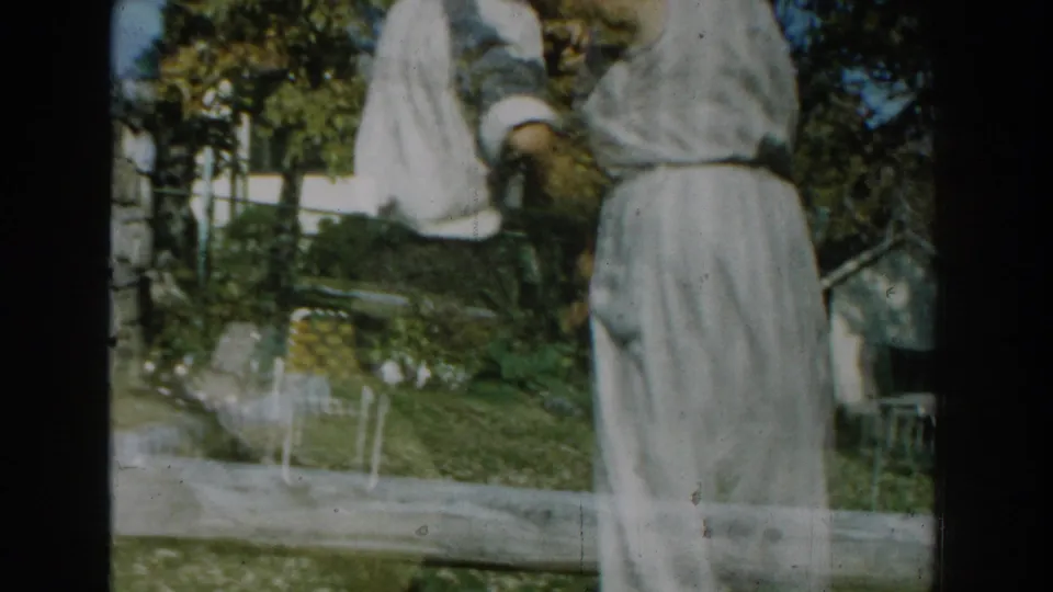 The elderly couple dances on the grass, with lawn chairs, shrubs, trees, and nearby houses visible behind them.