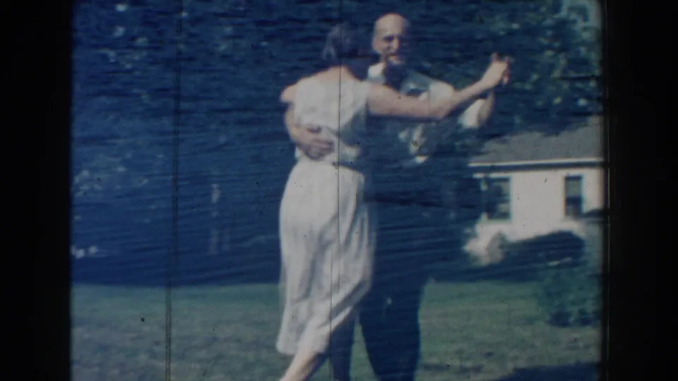 The dancing couple briefly faces the camera in this 1959 home-movie frame, with lawn, trees, and a house visible behind them.