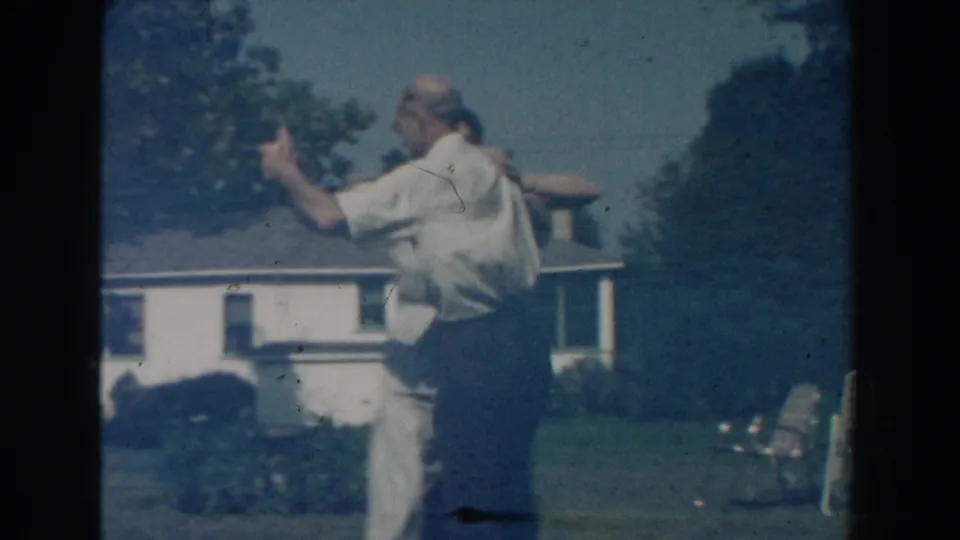 The man raises his hand mid-dance as the woman stays close, with the backyard and house still in view.